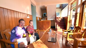 Dos personas sentadas en una mesa de madera con una computadora portátil, rodeadas de sillas de madera en una habitación con paneles de madera y una escalera al fondo.