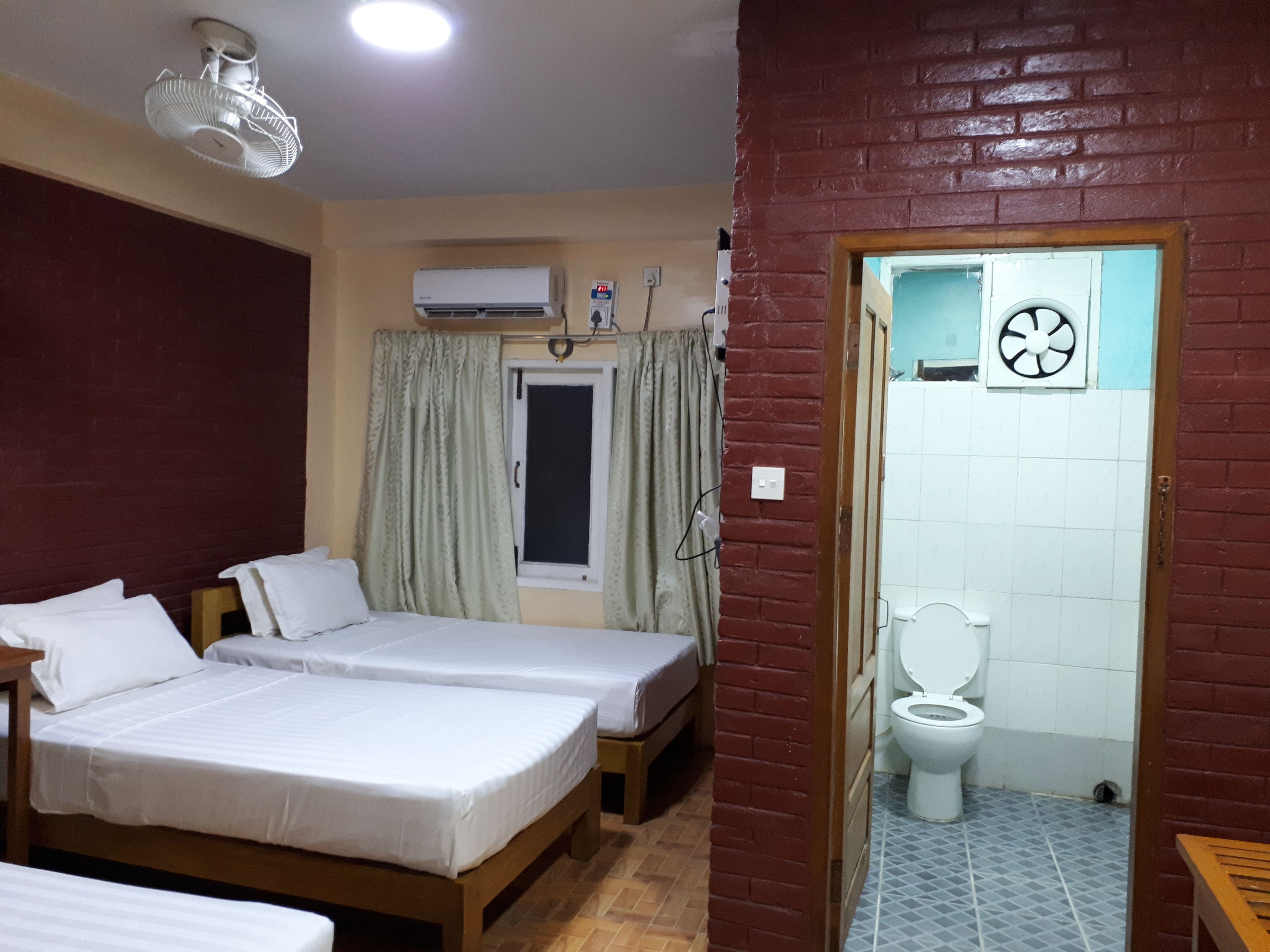 Una habitación con dos camas, una ventana con cortinas, un aire acondicionado, un inodoro y un banco de madera.