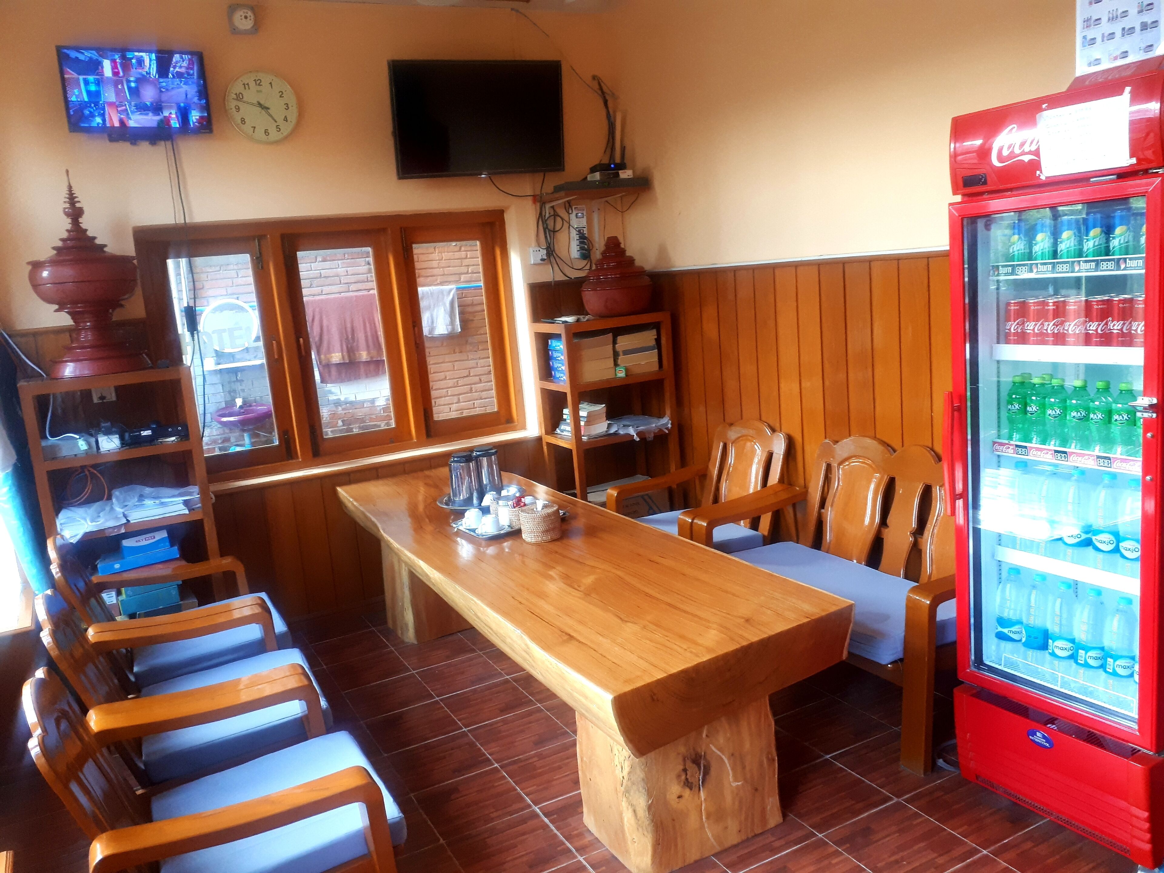 Een houten eettafel met stoelen, een aan de muur bevestigde televisie, een klok en een Coca-Cola koelkast.