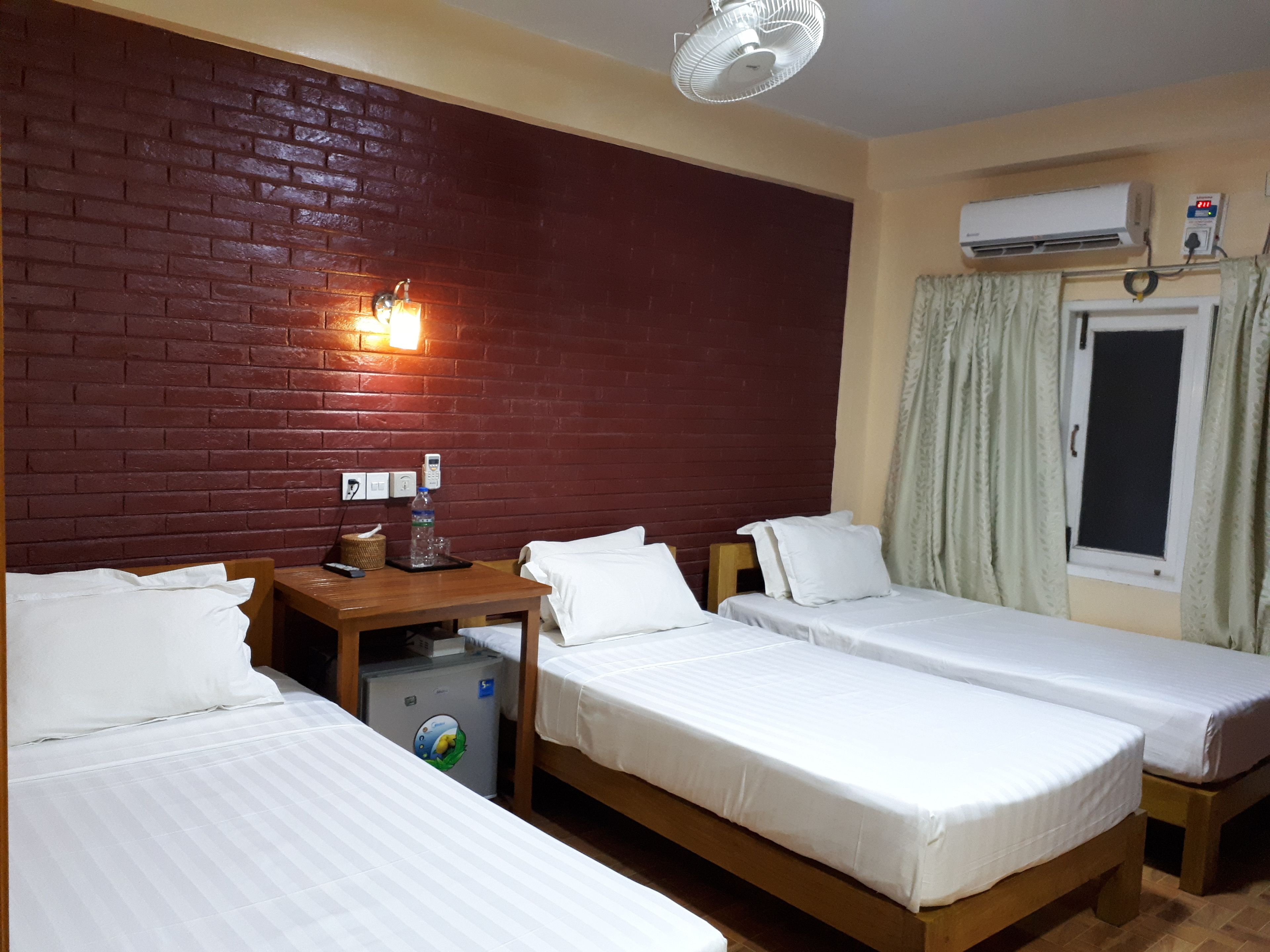 A hotel room with three single beds, a small table with a bottle and glasses, a wall-mounted air conditioner, and a window with curtains.