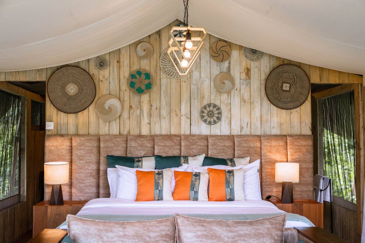 A bedroom with a wooden headboard, a bed with multiple pillows, two bedside tables with lamps, and wall decor.