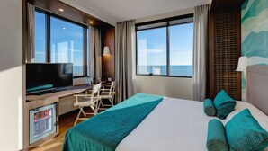 Habitación de hotel con una cama grande, un escritorio con televisor y vista al mar.