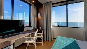 Habitación de hotel moderna con televisor grande, escritorio con sillas, cama y ventanas amplias con vista al mar.