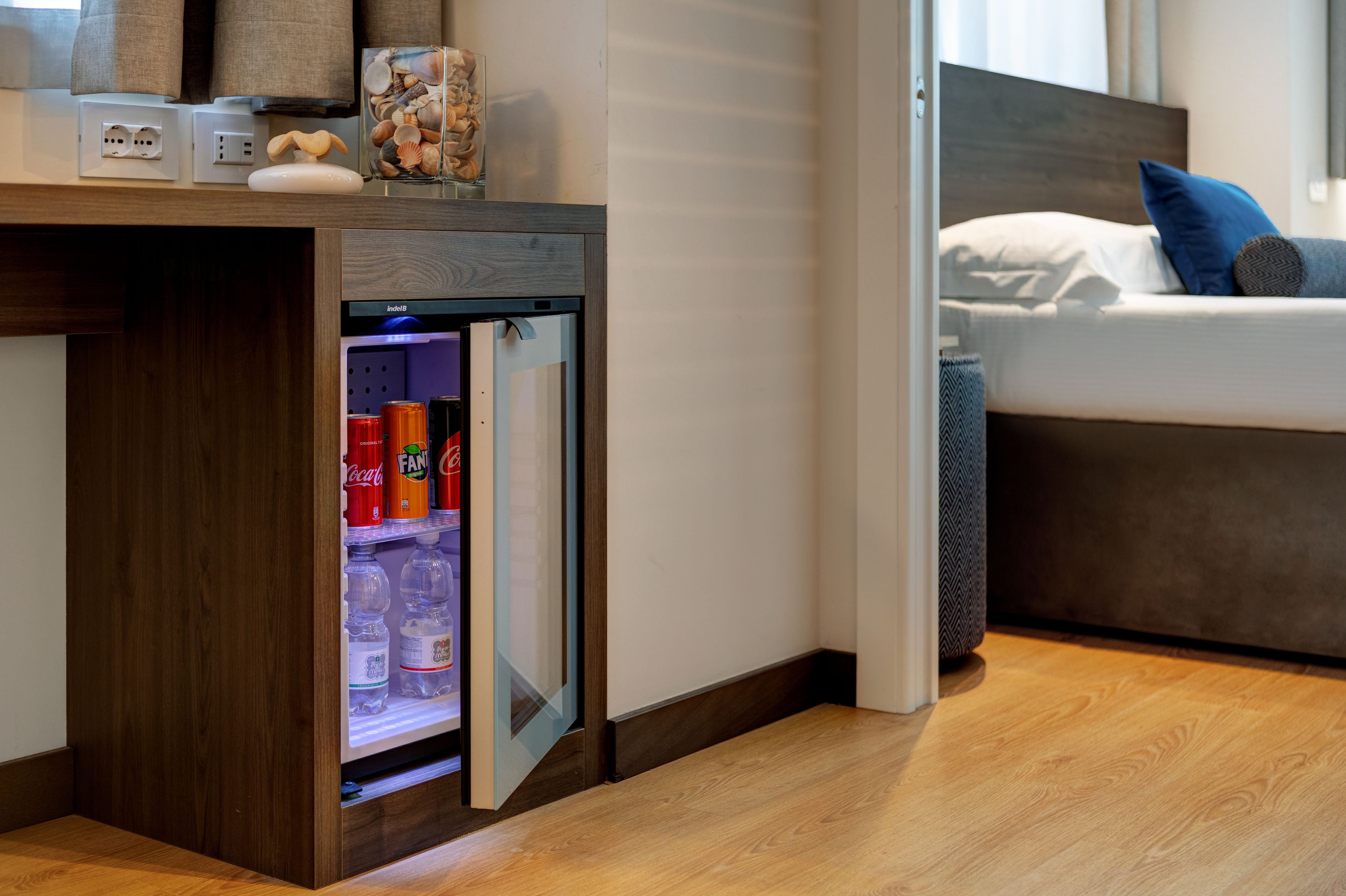 A compact refrigerator with beverages, a bedside table with a lamp, and a bed with pillows.