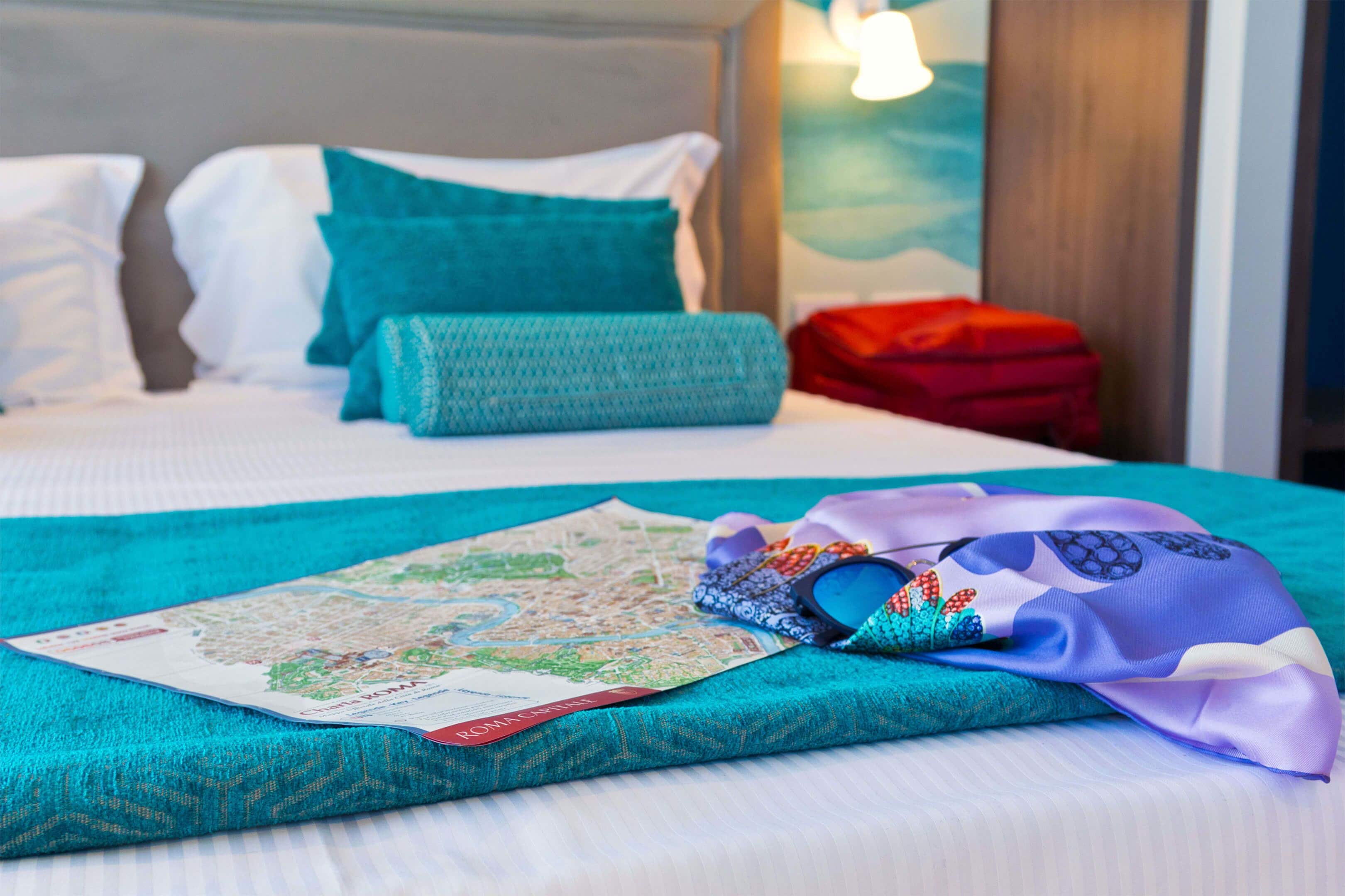 A neatly made bed with a map, a folded scarf, and a red bag on a teal bedspread.