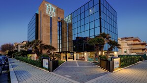 Un hotel moderno con fachada de vidrio, torre de ladrillo y un área de entrada pavimentada con palmeras.