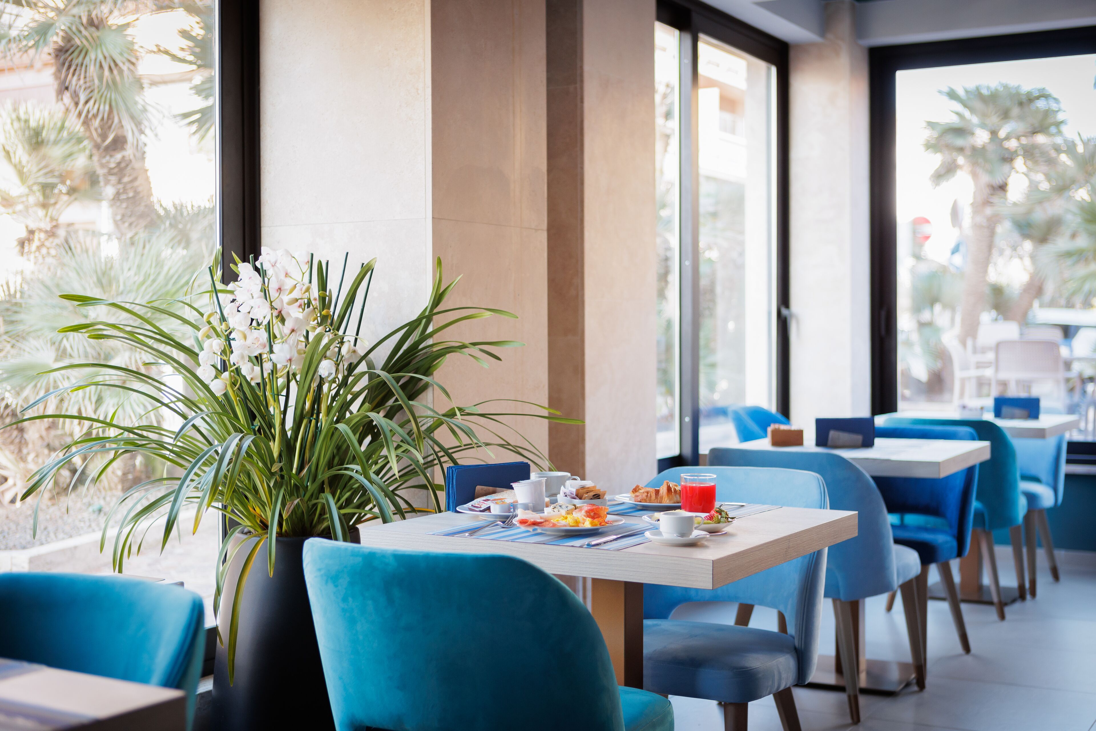 Uma área de refeições moderna com cadeiras azuis, uma mesa posta para o café da manhã e uma grande janela com vista para palmeiras.