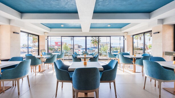 Un comedor espacioso con sillas azules y mesas blancas, amplias ventanas que ofrecen vistas al exterior.