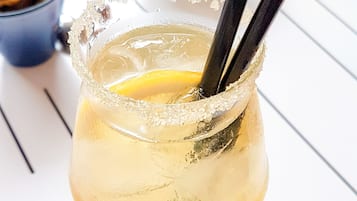 Un vaso de cóctel con borde salado y una rodaja de limón, acompañado de un paño de papel azul y una taza de metal al fondo.