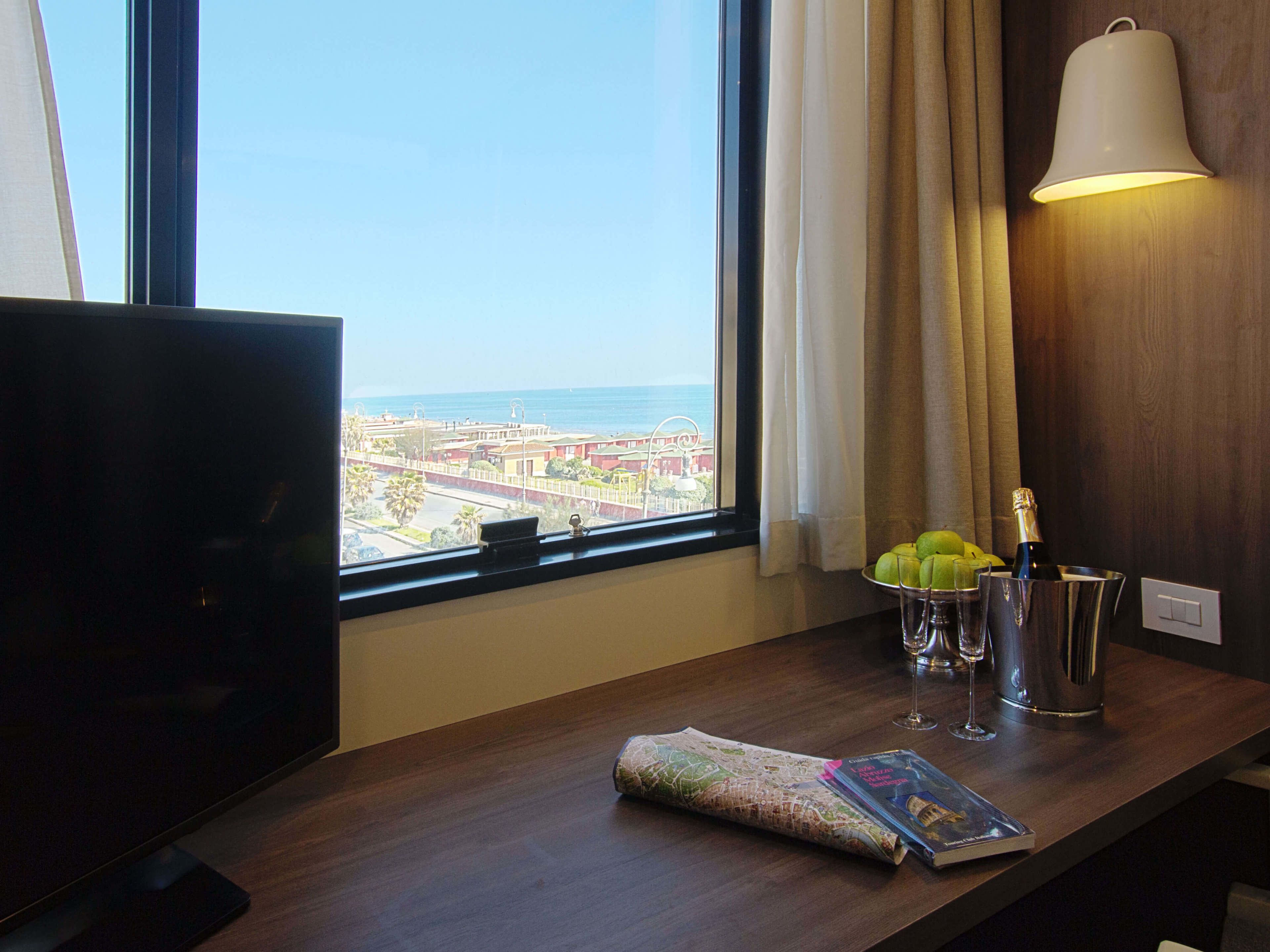 A hotel room with a TV, a table with a map, a bottle of champagne, and a view of the city and ocean.