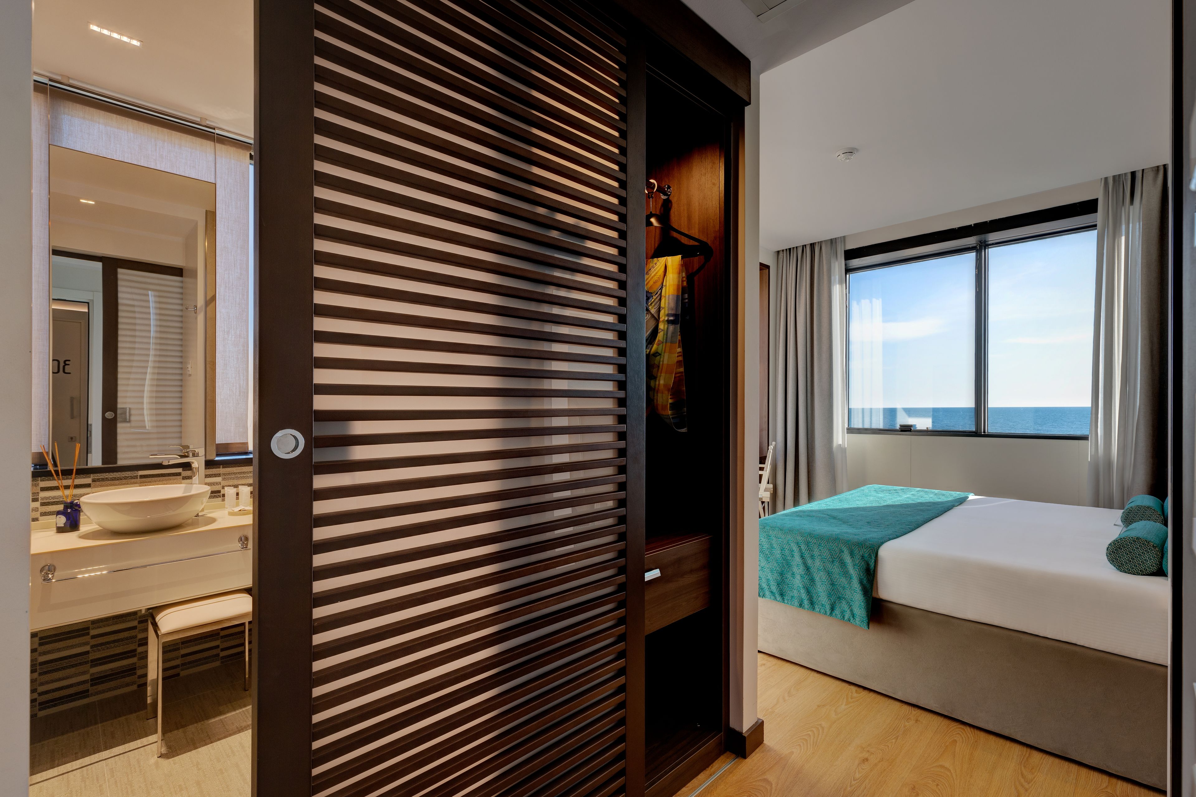 A modern hotel room with a large window, a bed, a bathroom with a sink, and a wardrobe.