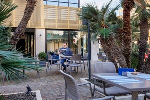 An outdoor seating area with tables and chairs, surrounded by palm trees and a building with a modern design.