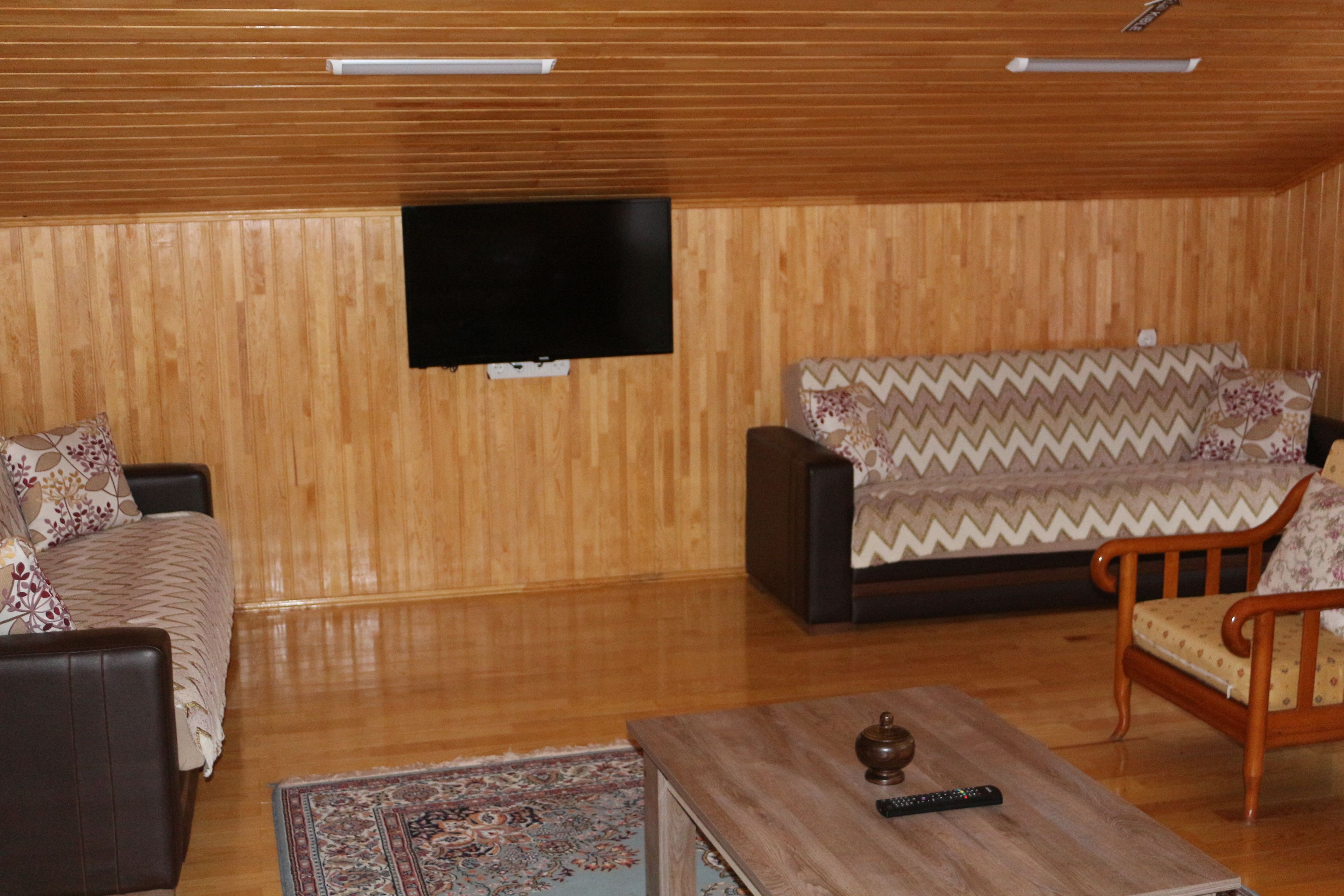 A living room with a wooden paneled ceiling, a flat-screen TV mounted on the wall, a sofa with a patterned cover, a wooden coffee table with a remote control, and a patterned rug on the floor.