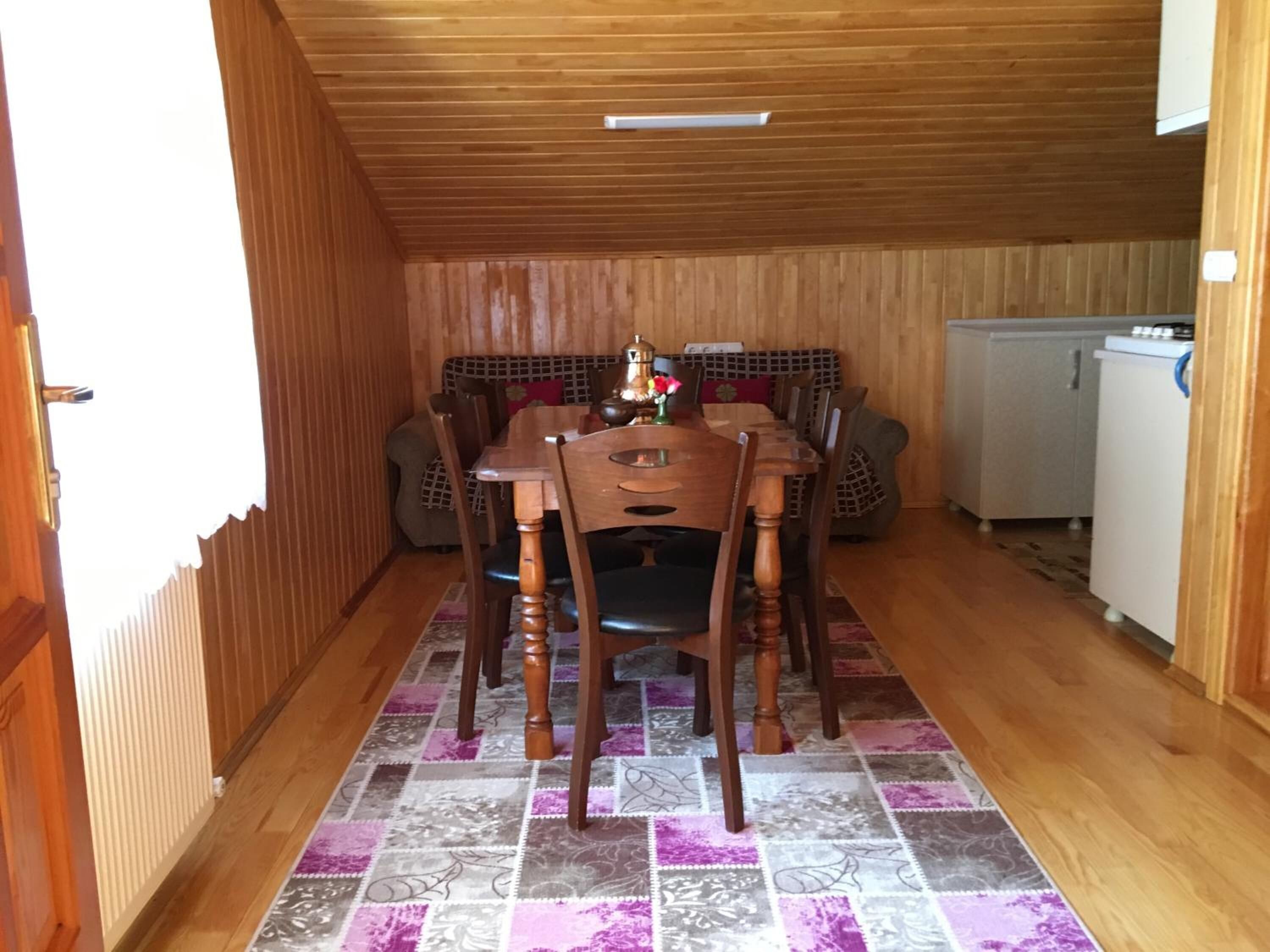 A wooden-paneled room with a dining table and chairs, a kitchen area with a refrigerator, and a patterned rug on the floor.