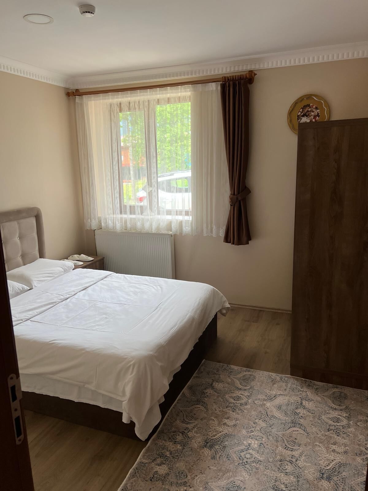 A bedroom with a bed, a nightstand, a window with curtains, a radiator, and a wooden wardrobe.