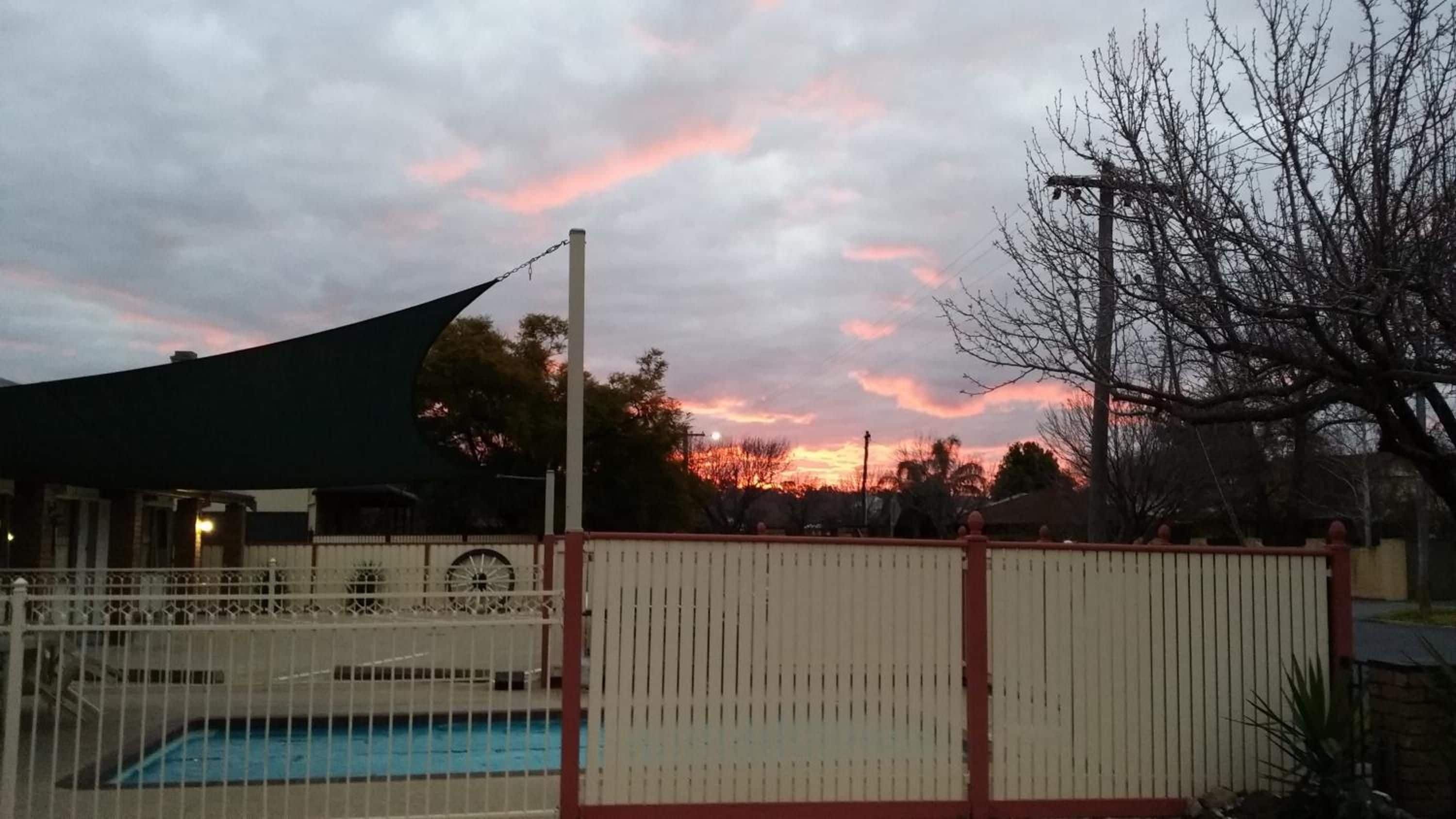 A swimming pool with a fence, a building with a dark awning, and a sunset sky.