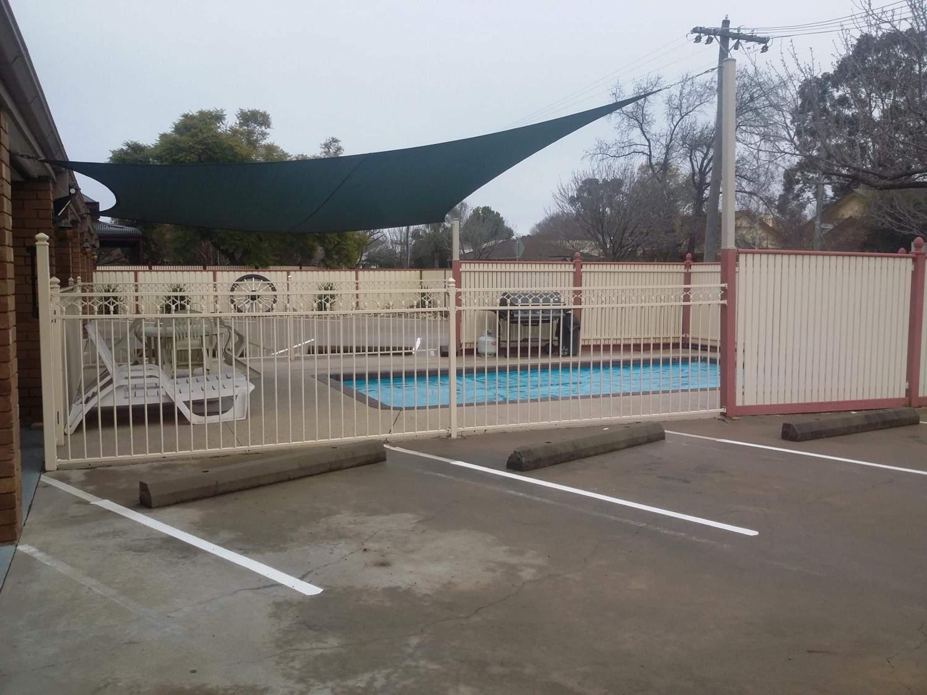 A swimming pool with a white fence, a shaded area, and a brick building in the background.