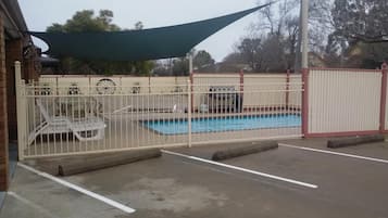 A swimming pool with a white fence, a shaded area, and a brick building in the background.