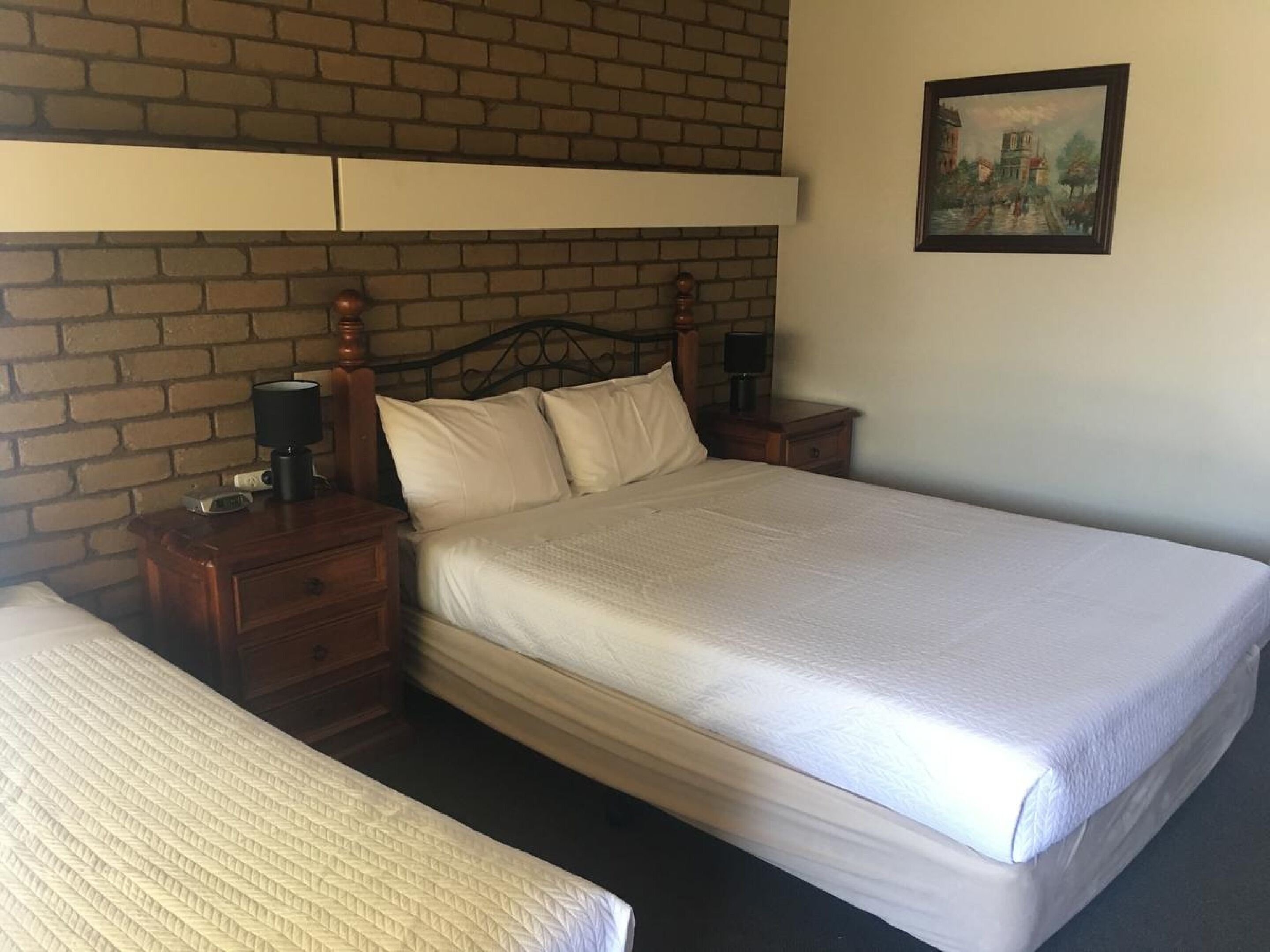Una cama doble con cabecera de madera, una mesita de noche con lámpara, y un cuadro en la pared.