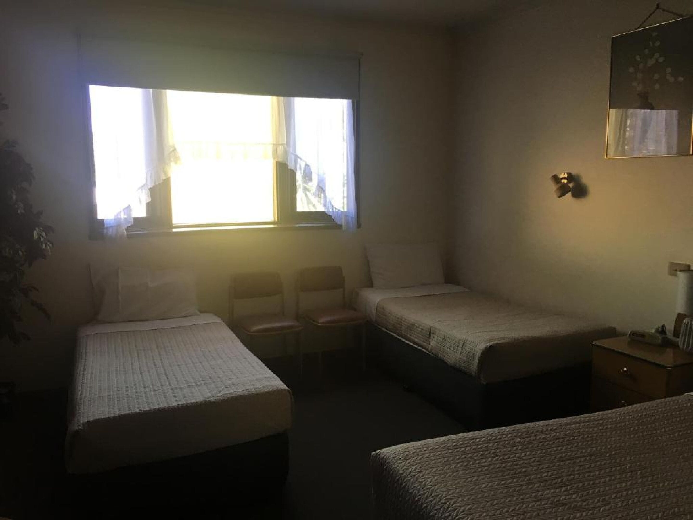 A small, dimly lit room with two single beds, a small table, and a chair. There is a window with curtains, a plant, and a wall-mounted light fixture.