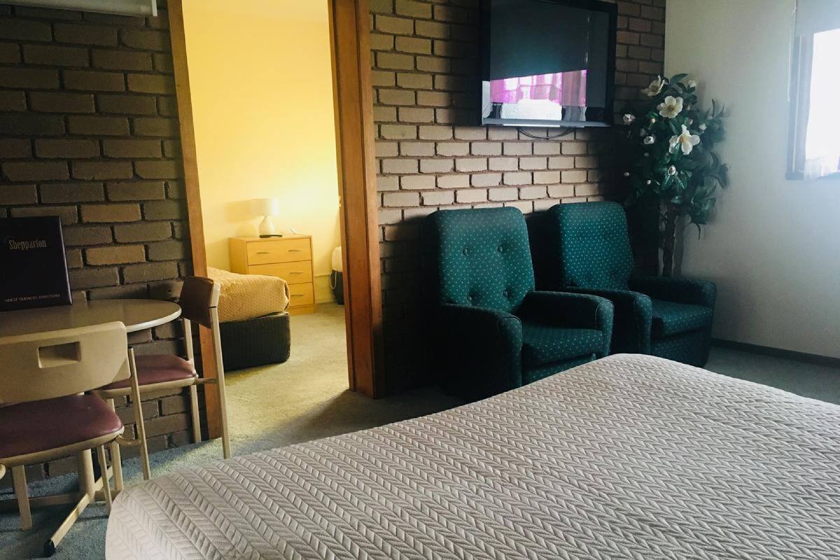 Habitación de hotel con una cama grande, un escritorio con una silla, dos sillones verdes, un aparador pequeño de madera, un televisor montado en una pared de ladrillo y una planta en maceta.