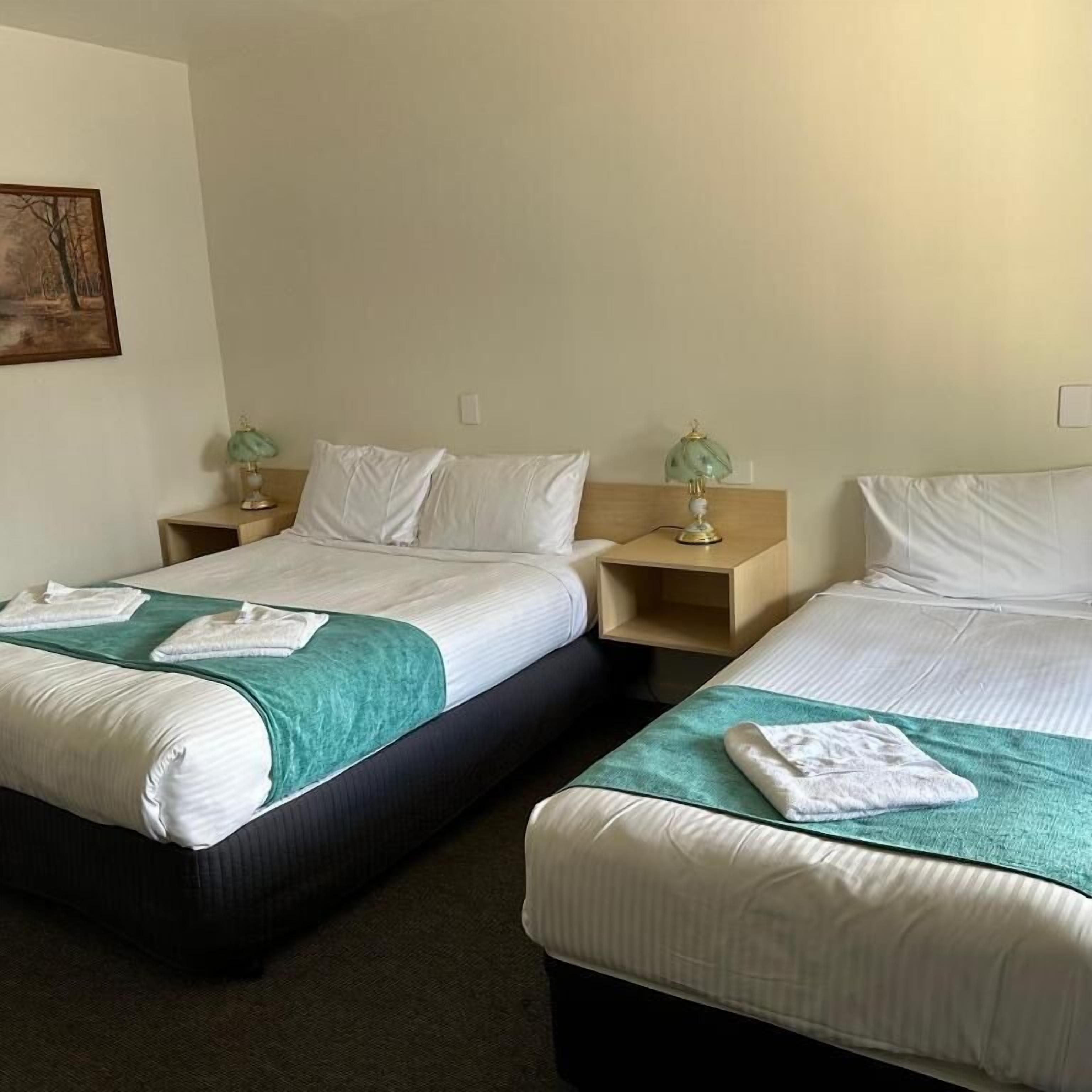 A hotel room with two beds, each with a green blanket and white pillows. There are bedside tables with lamps and a painting on the wall.