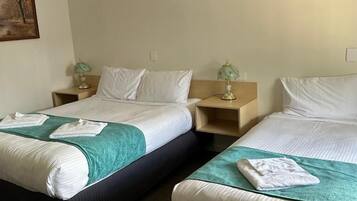 A hotel room with two beds, each with a green blanket and white pillows. There are bedside tables with lamps and a painting on the wall.