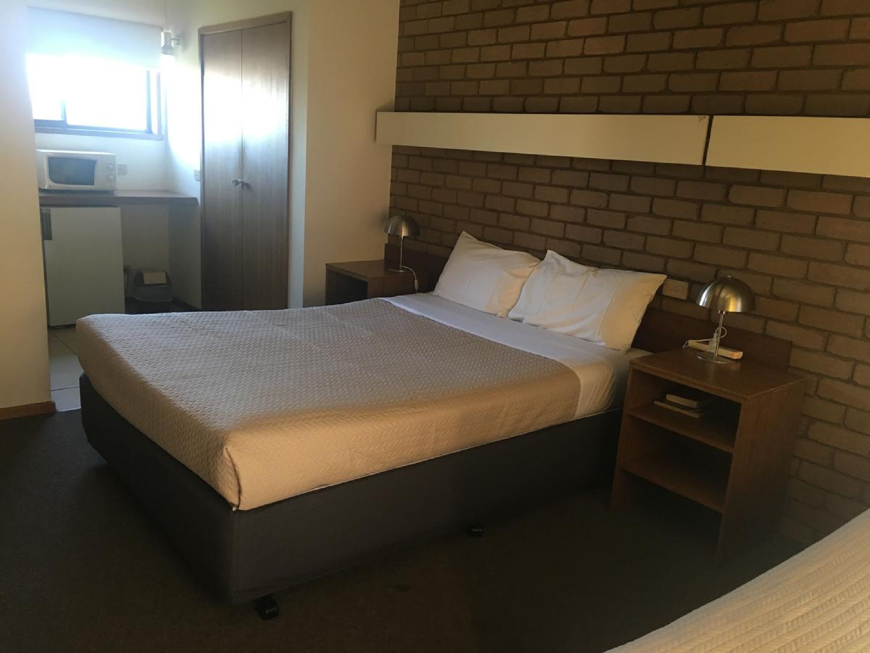 A hotel room with a bed, two bedside tables, a lamp, a small desk with a chair, a microwave, and a wardrobe.