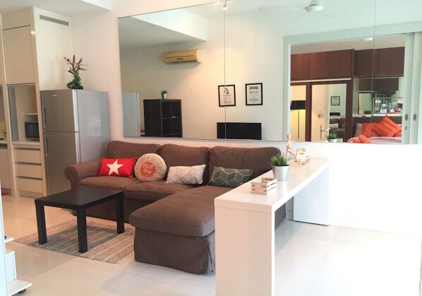 A modern living room with a sectional sofa, a small coffee table, and a white console table. There is a kitchen area visible through an open door. - Marc Residence B2906 by SYNC (Kuala Lumpur)