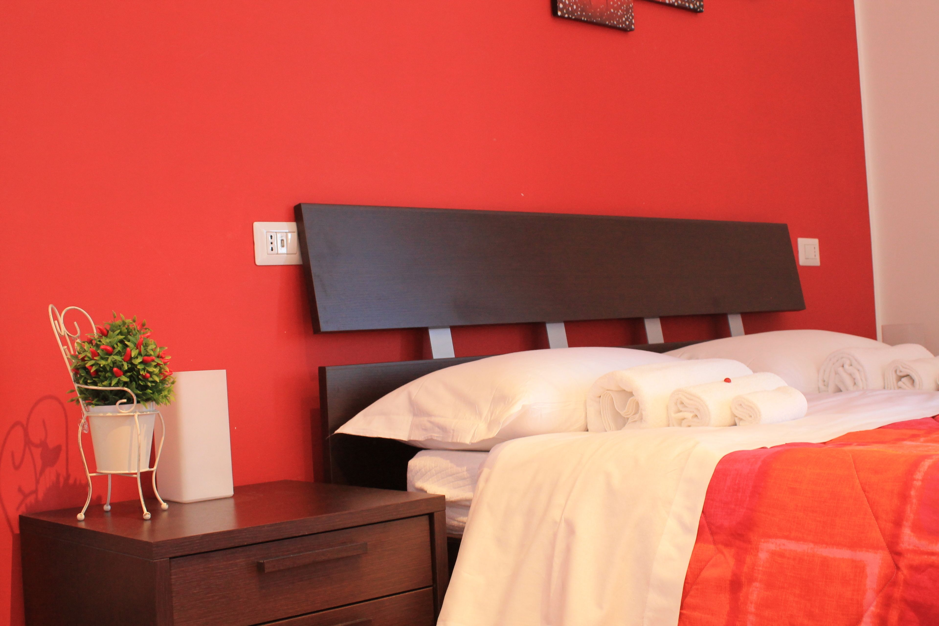Una cama bien hecha con sábanas blancas y una manta roja, una mesita de noche con una lámpara blanca y una planta en maceta, y un cabecero de madera oscura.