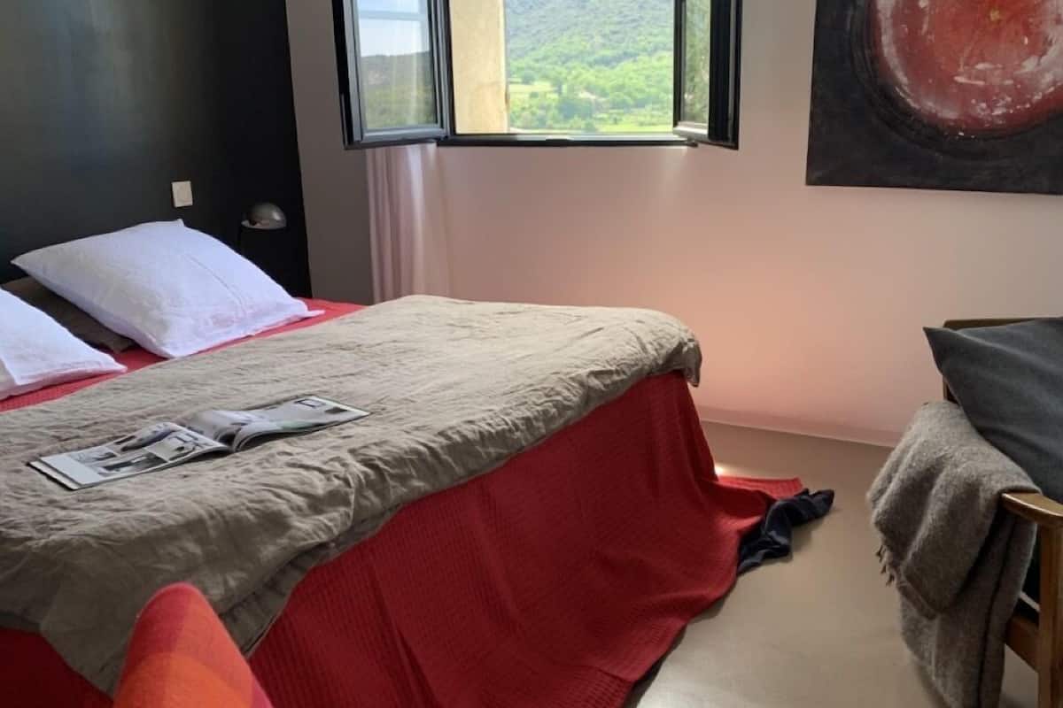 Quarto com cama, uma cadeira e janela com vista para vegetação.