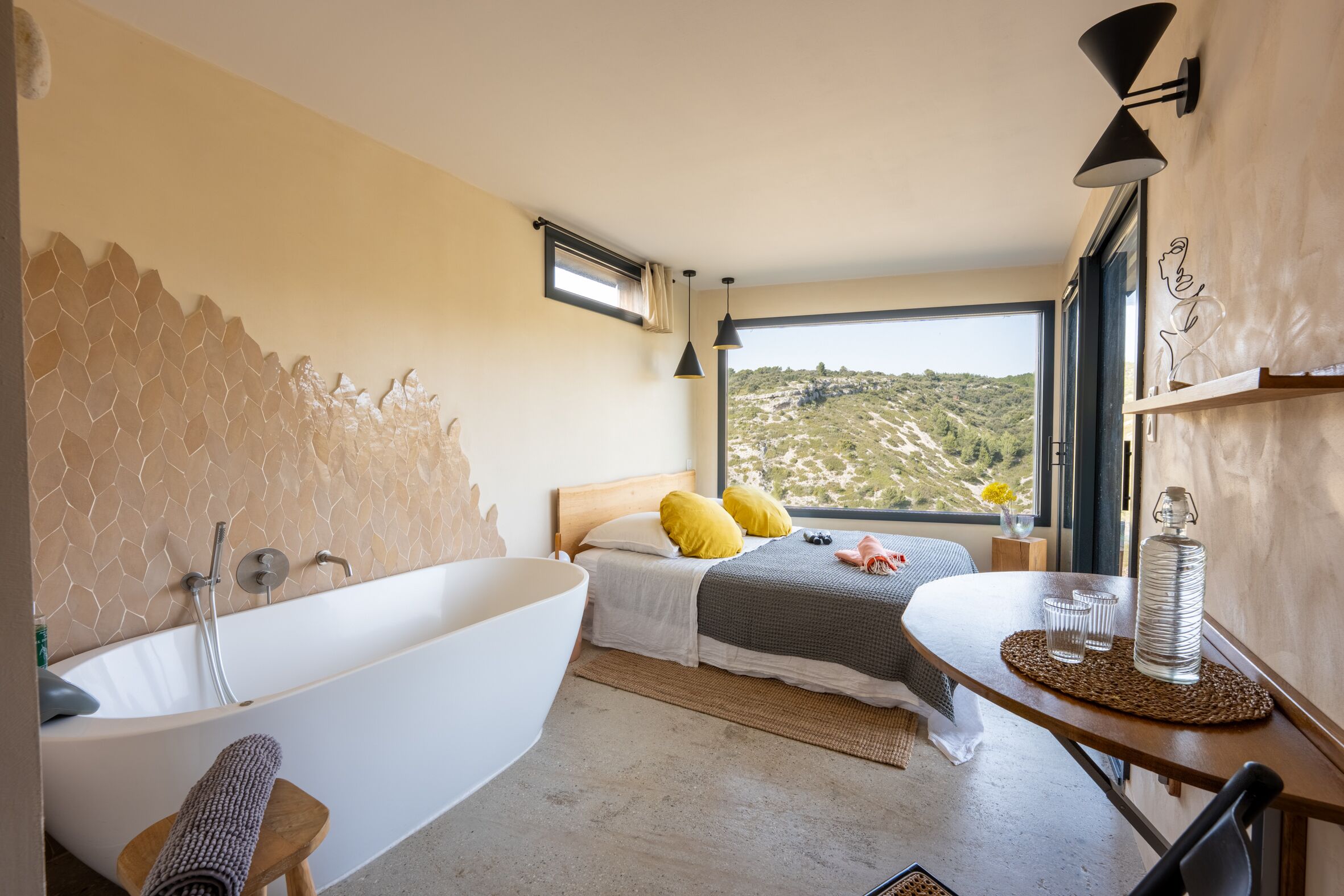 A modern bedroom with a large bathtub, a bed with yellow pillows, a view of a hilly landscape, and a small table with glasses and a bottle.
