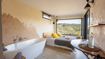 A modern bedroom with a large bathtub, a bed with yellow pillows, a view of a hilly landscape, and a small table with glasses and a bottle.