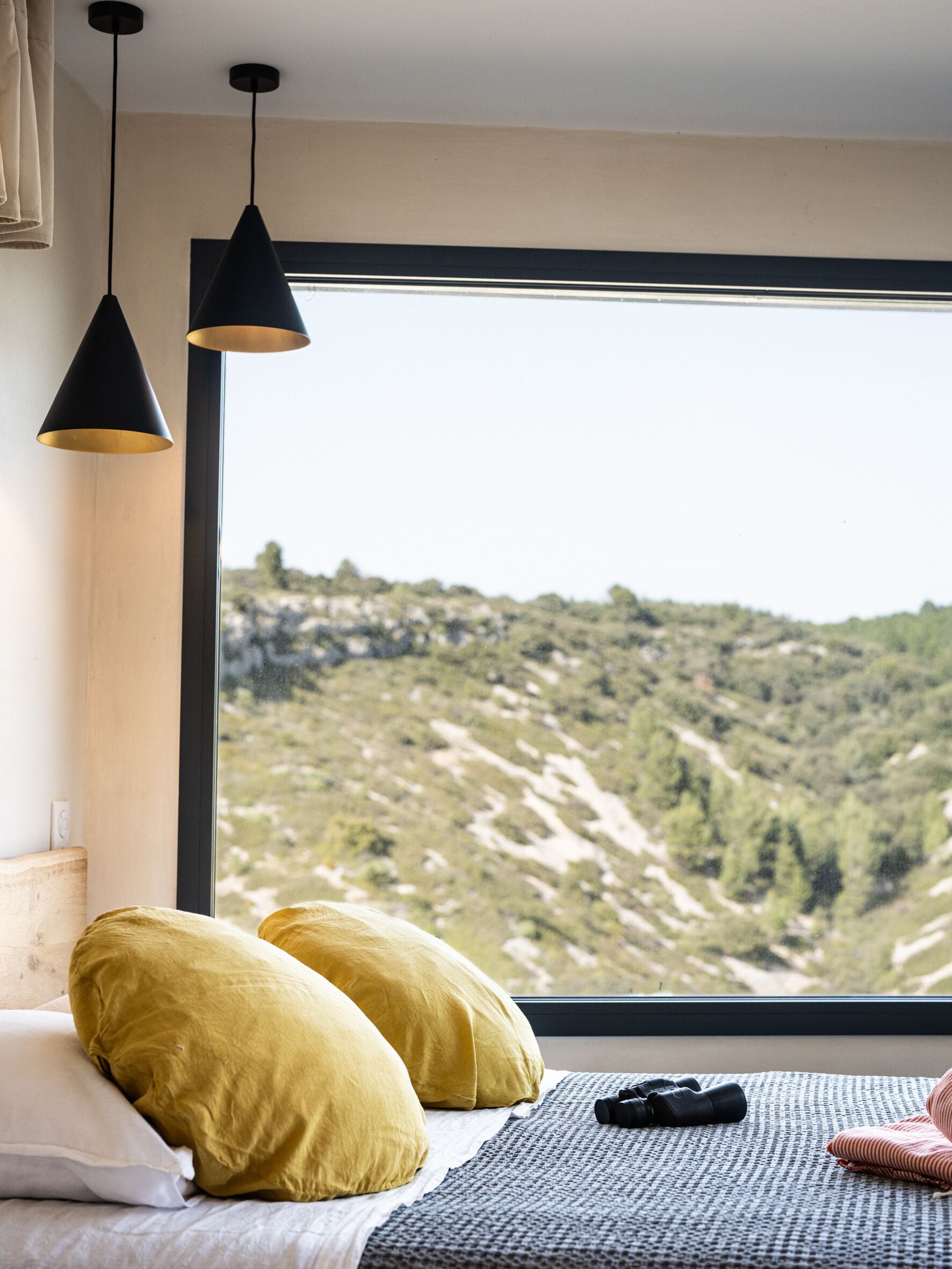 A bedroom with a large window, two pillows, a bed, and a pair of binoculars.