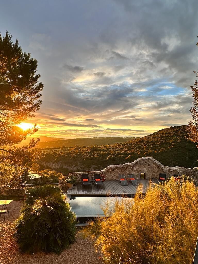 A rustic stone building with a sloped roof, surrounded by trees and a view of a hilly landscape.