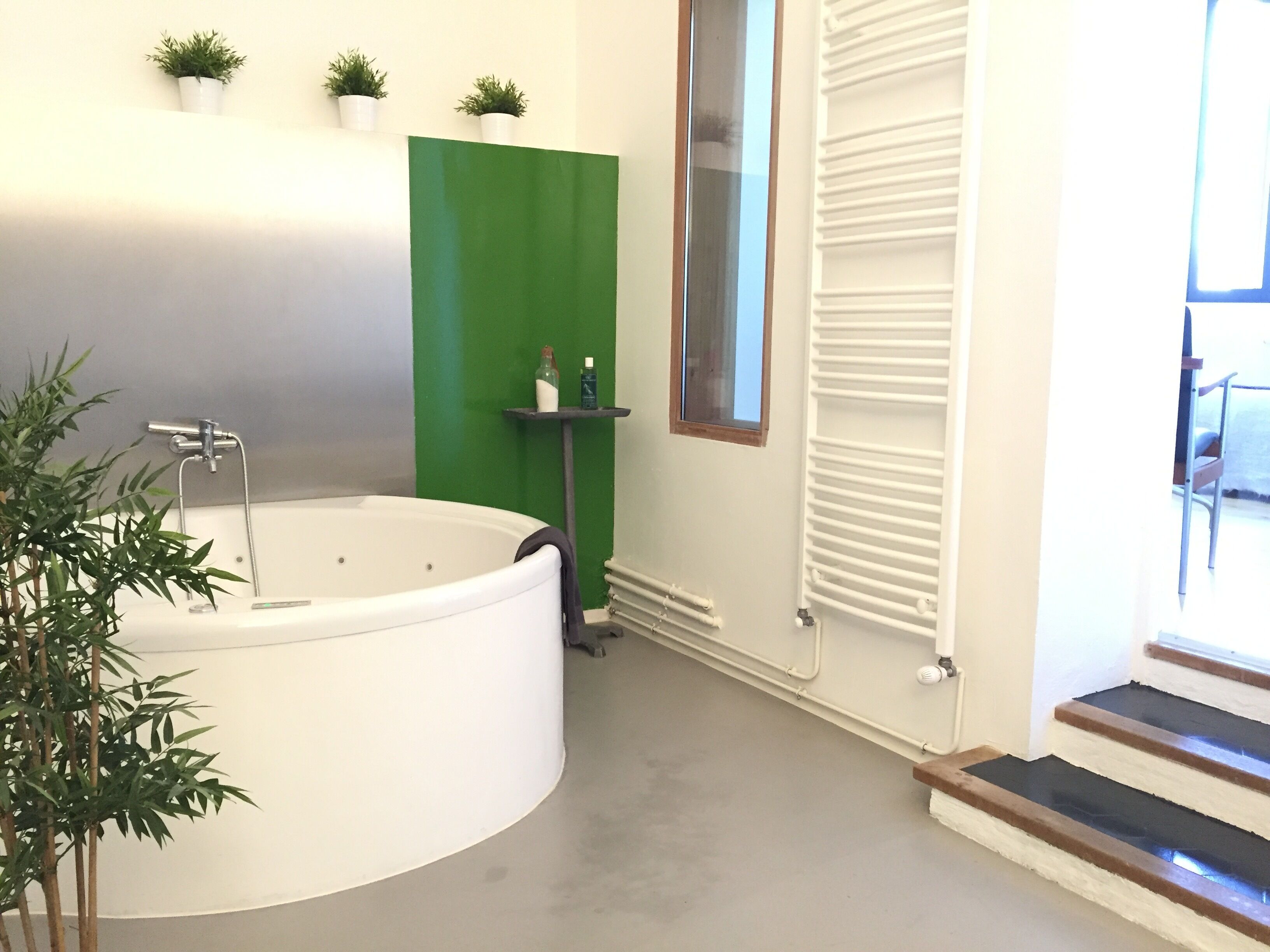 A modern bathroom with a large bathtub, a green accent wall, a small window, and a radiator.