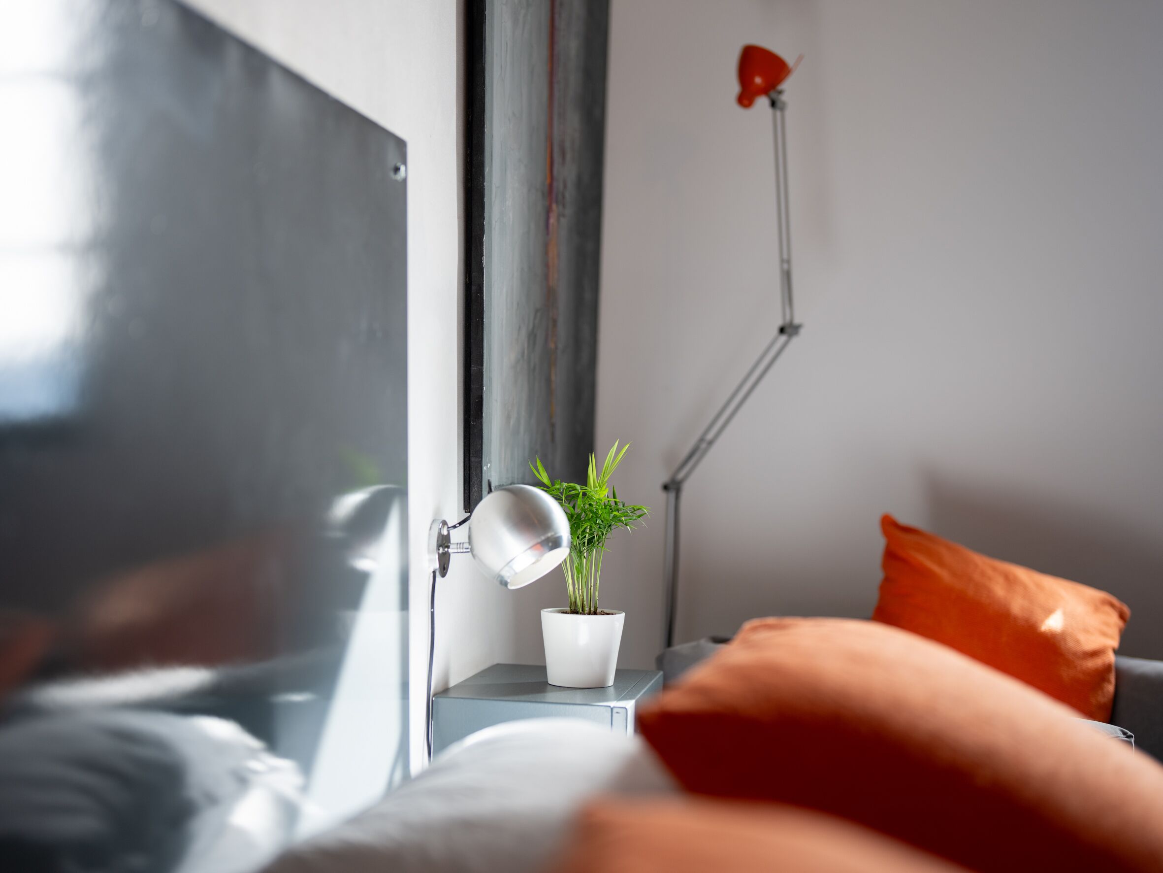 Una cama con un almohadón blanco, uno rojo y una pequeña planta en maceta sobre una mesita de noche.