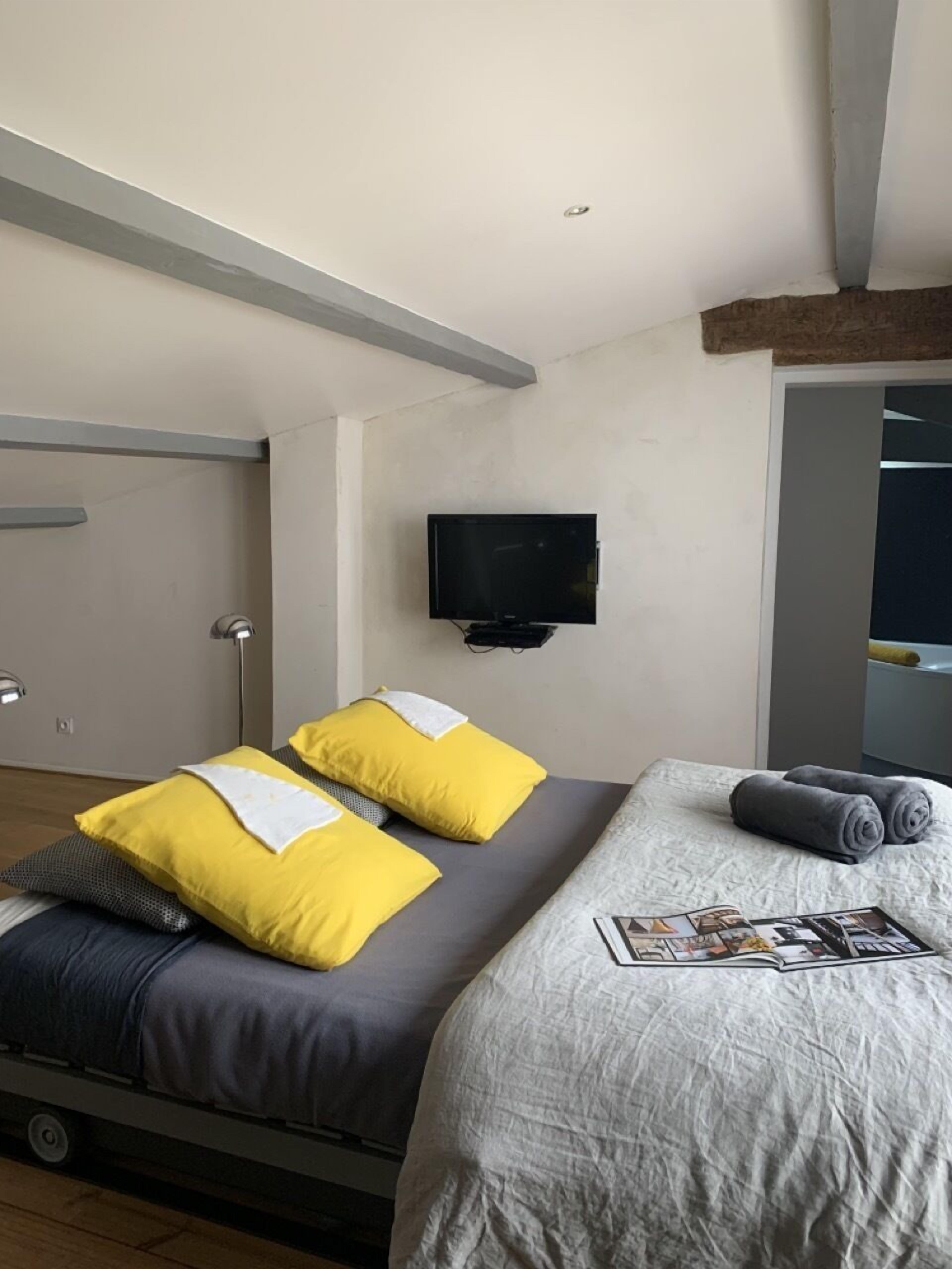 A modern hotel room with a bed, a flat-screen TV mounted on the wall, and a wooden ceiling beam.