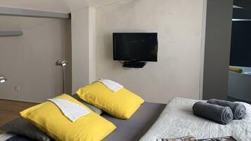 A modern hotel room with a bed, a flat-screen TV mounted on the wall, and a wooden ceiling beam.