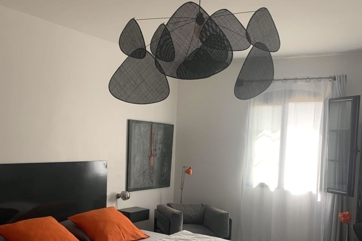 Quarto moderno com cama grande, duas almofadas laranja, luminária de teto preta e sofá cinza, além de janela com cortinas leves.