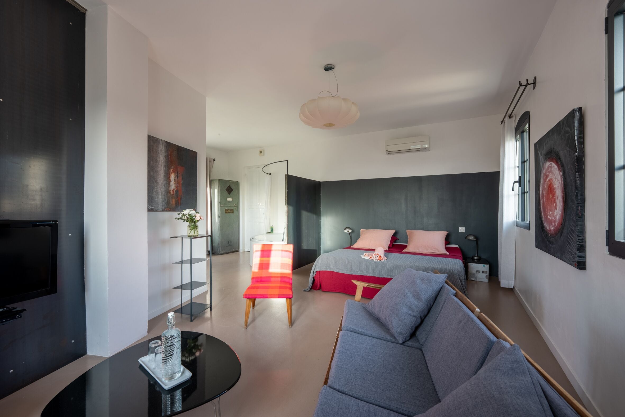 Una habitación de hotel moderna con una televisión de pantalla plana, un sofá, una cama con ropa de cama roja y obras de arte en las paredes.