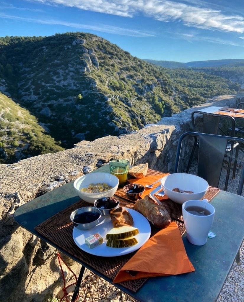 Une table dressée avec des aliments pour le petit-déjeuner, notamment du pain, des fruits et des boissons, donnant sur un paysage montagneux.