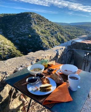 A table set with breakfast items, including bread, fruit, and beverages, overlooking a mountainous landscape. - Metafort (Méthamis)