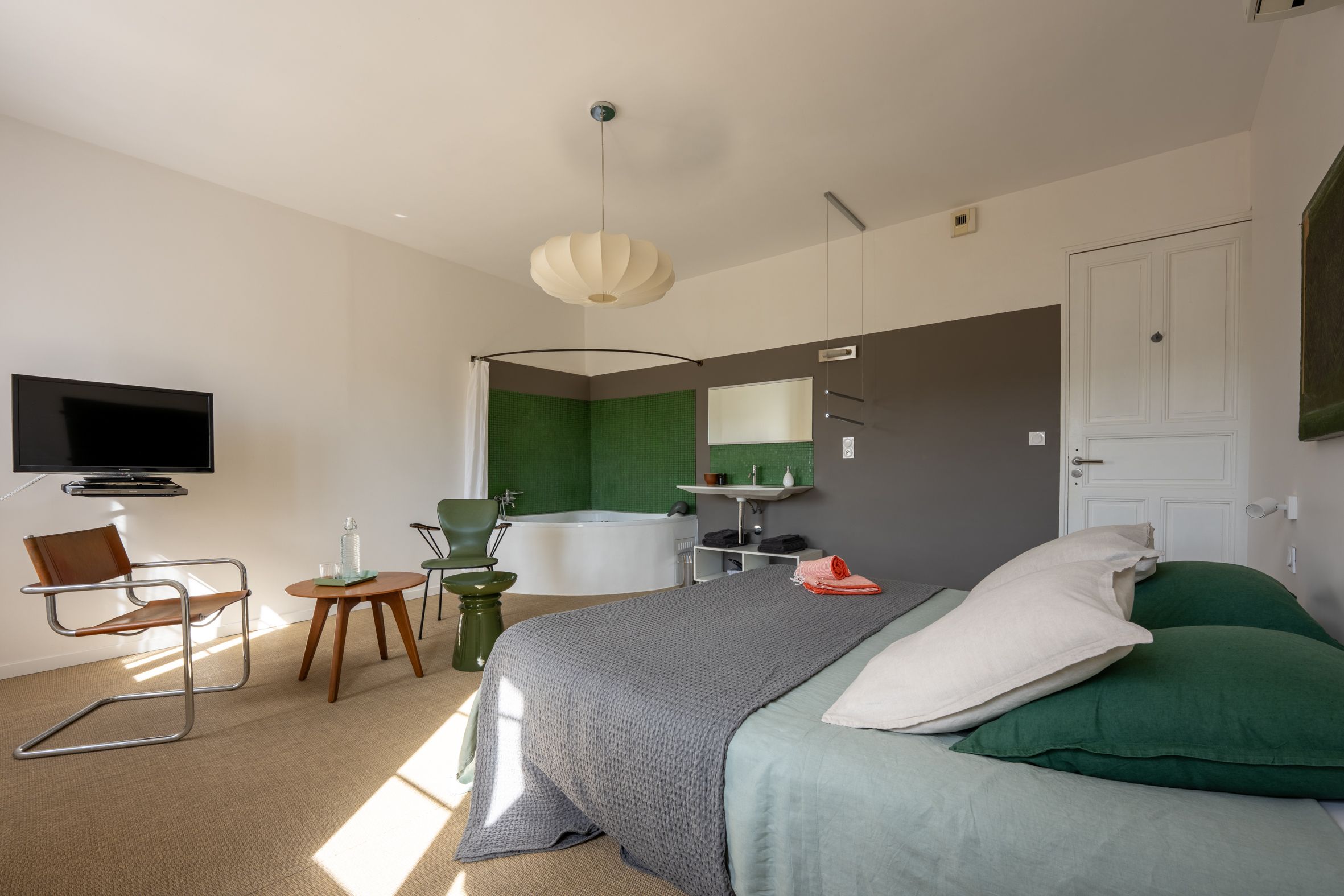 Een moderne slaapkamer met een bed, een televisie, een stoel, een tafel en een badkamer met een badkuip en groene tegels.