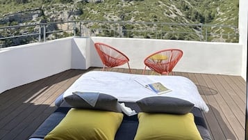 A balcony with a bed, two red chairs, and a view of a hilly landscape.