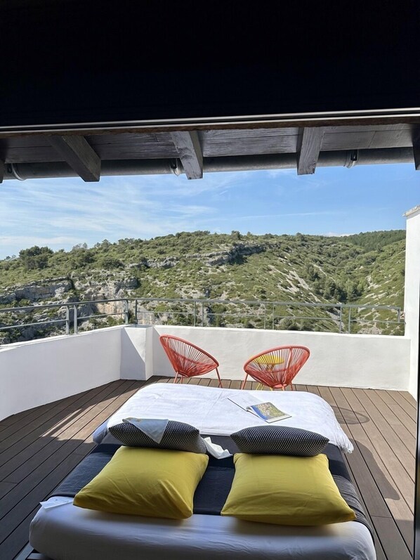 A balcony with a bed, two red chairs, and a view of a hilly landscape.