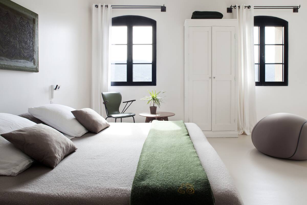 Quarto de hotel moderno com uma cama grande, uma mesinha com uma planta, uma cadeira verde e um banquinho redondo.