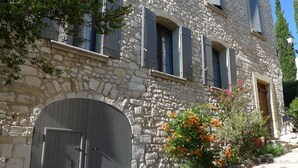 A stone building with a large wooden door, multiple windows, and a small garden with orange flowers. - Metafort (Méthamis)