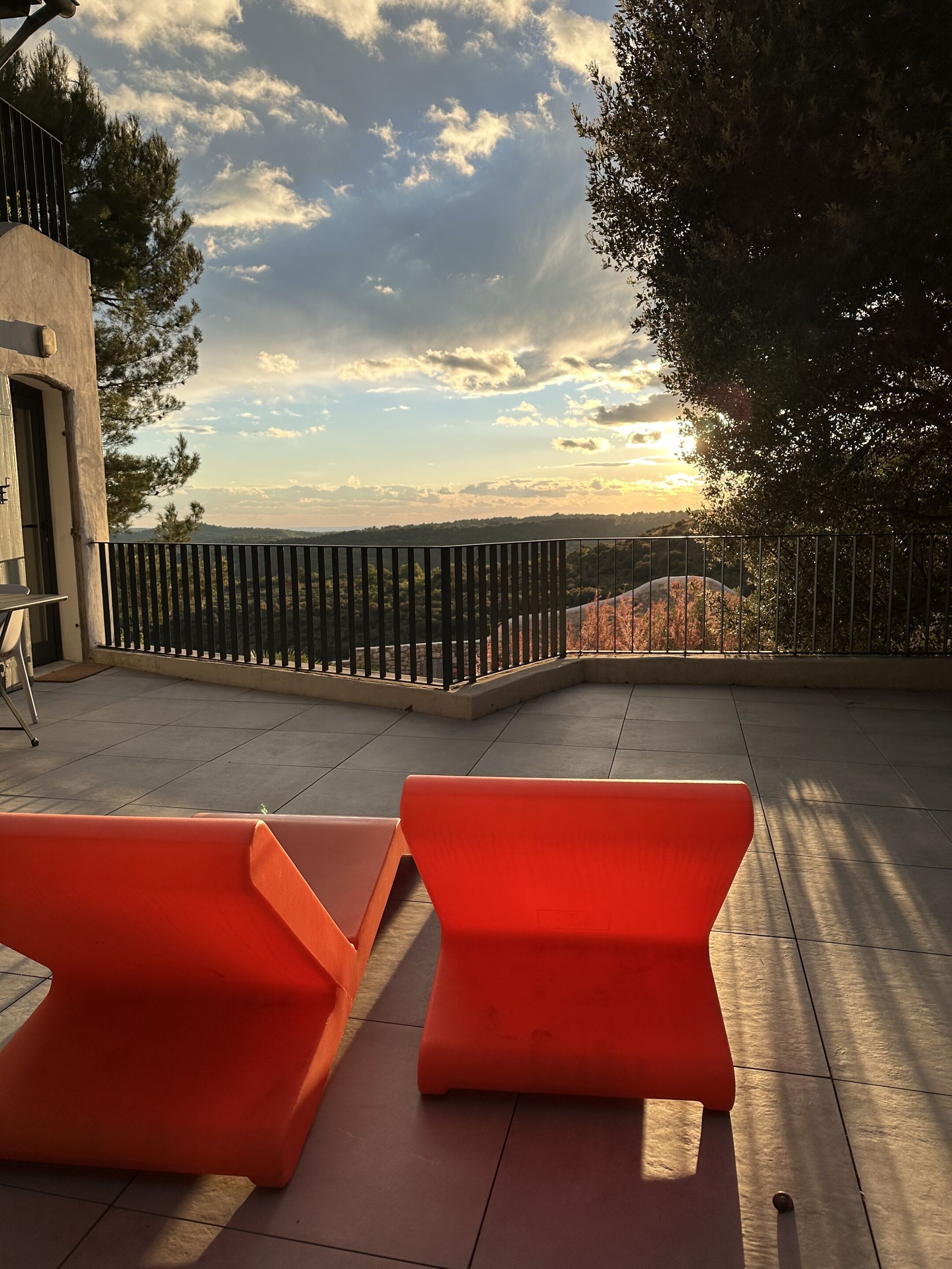 Une terrasse avec deux chaises orange, une rambarde en métal et une vue sur un paysage vallonné sous un ciel partiellement nuageux.
