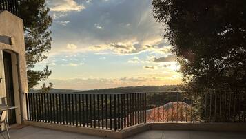 Une terrasse avec deux chaises orange, une rambarde en métal et une vue sur un paysage vallonné sous un ciel partiellement nuageux.